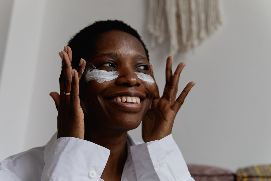 Black woman applying face cream indoors, smiling joyfully in natural lighting.