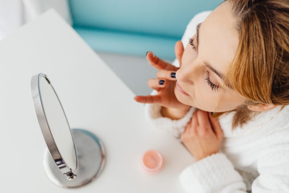 A woman in a white robe applies skincare cream while looking at herself in a mirror.