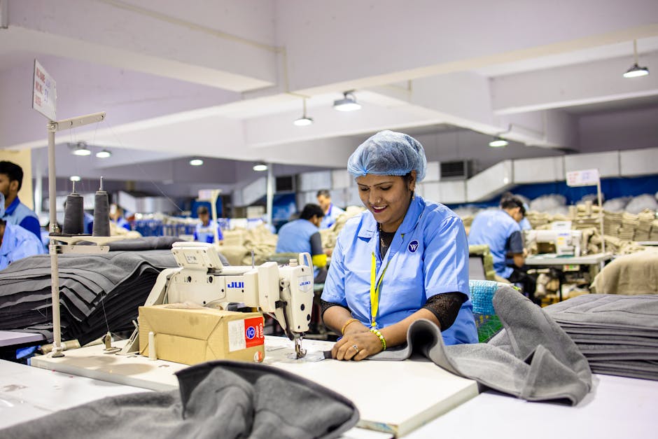Smiling factory workers operate sewing machines in a bustling textile factory environment.