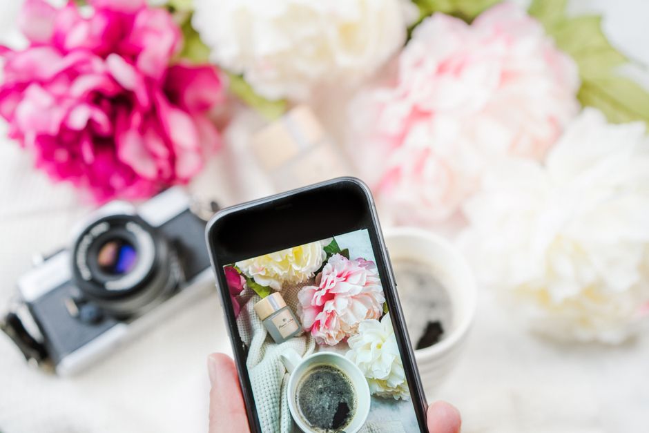 A smartphone captures a blooming floral arrangement with coffee and vintage camera.