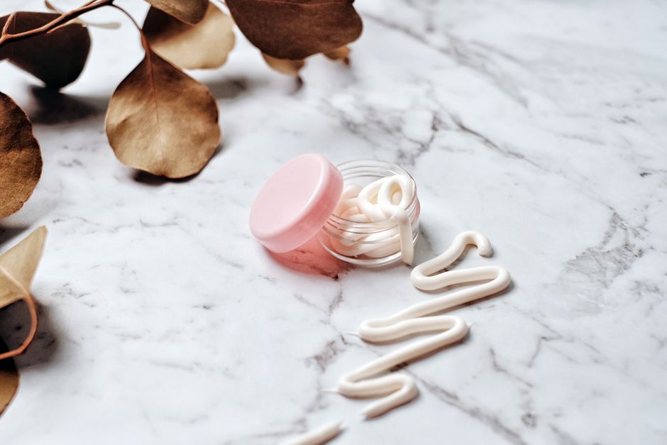 Close-up of skincare cream in a pink glass jar on a marble surface surrounded by leaves, i