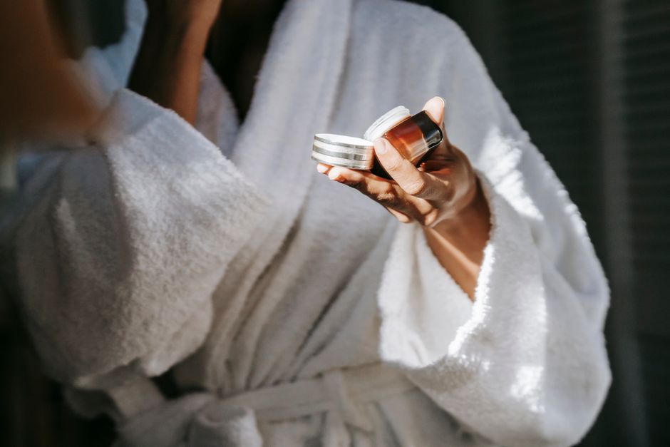 Crop anonymous ethnic female in bathrobe applying moisturizing cream on face while reflect