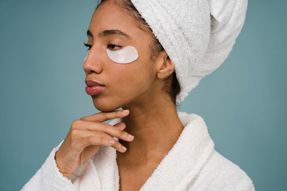 Pensive African American female with eye patches in white towel on head and bathrobe touching face and looking away against blue background