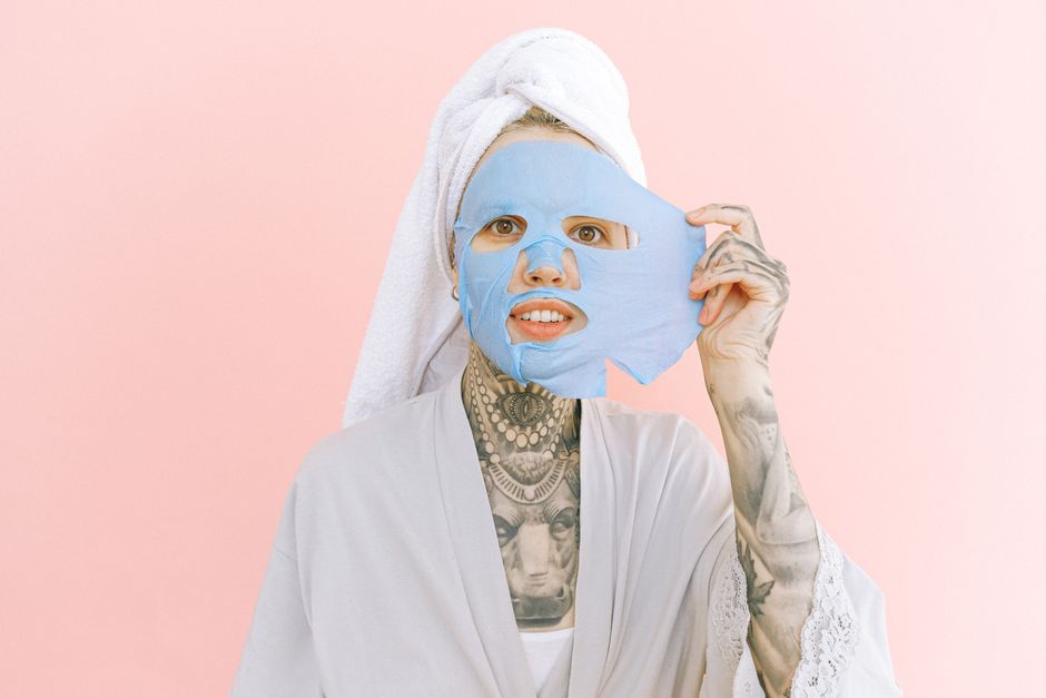 Cheerful young female in dressing gown and white terry towel on head taking off blue collagen face mask while standing on pale pink background and looking at camera