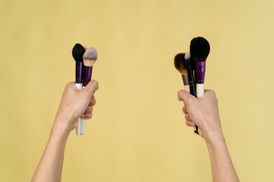 Two hands holding various makeup brushes against a yellow background.