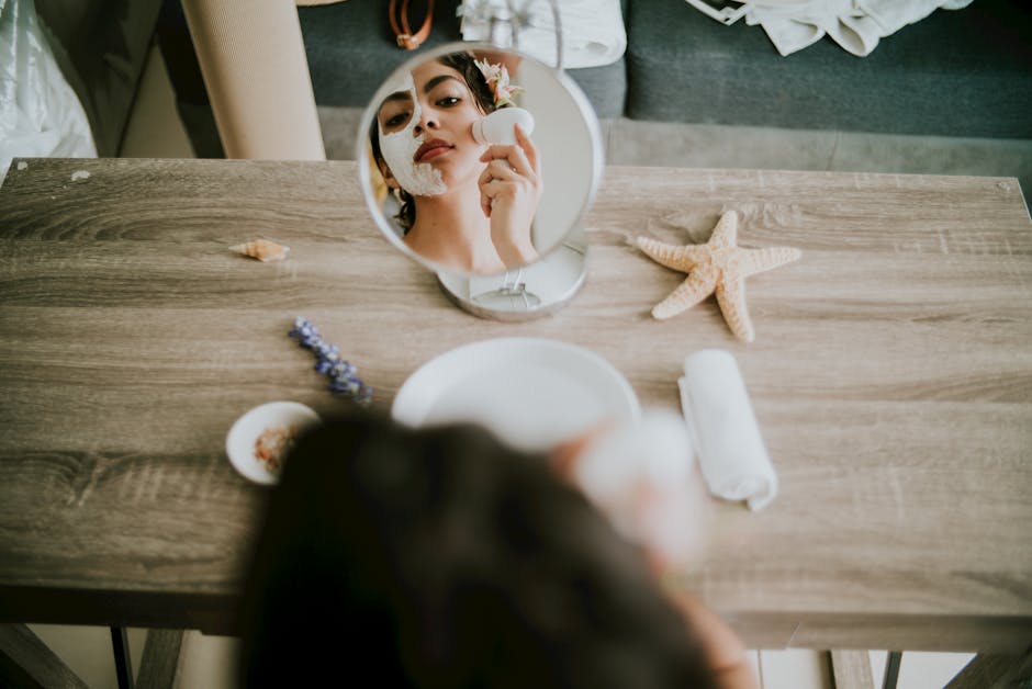 A woman applies a facial mask in front of a mirror with spa elements on a wooden table.