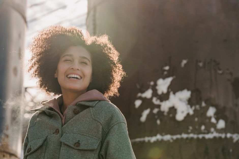 Happy young ethnic female with curly hair wearing warm clothes laughing merrily on street on sunny winter day