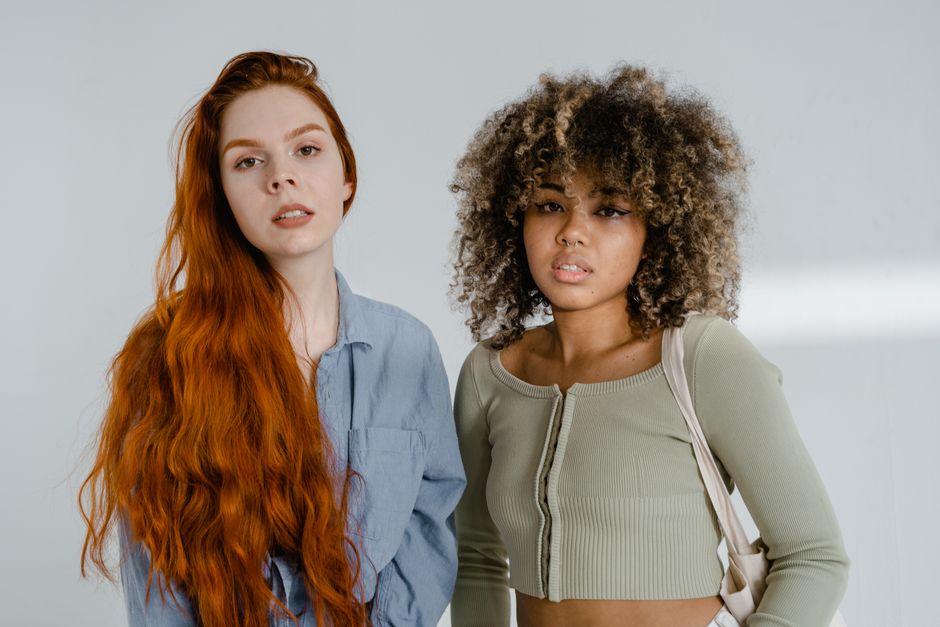 Portrait of two diverse women standing confidently indoors, showcasing unique hair styles.
