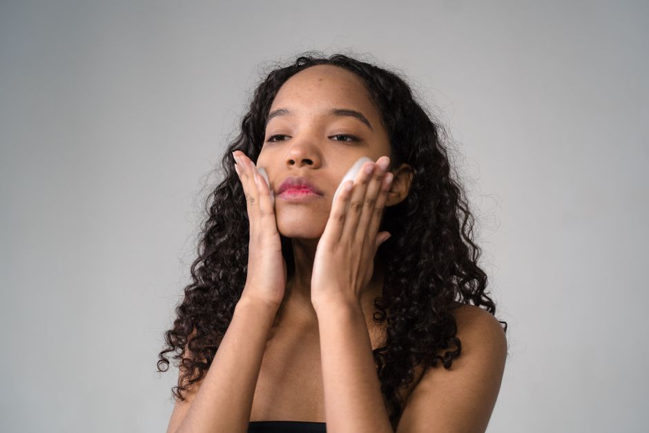 A young woman with curly hair gently applies skincare cream to her face.