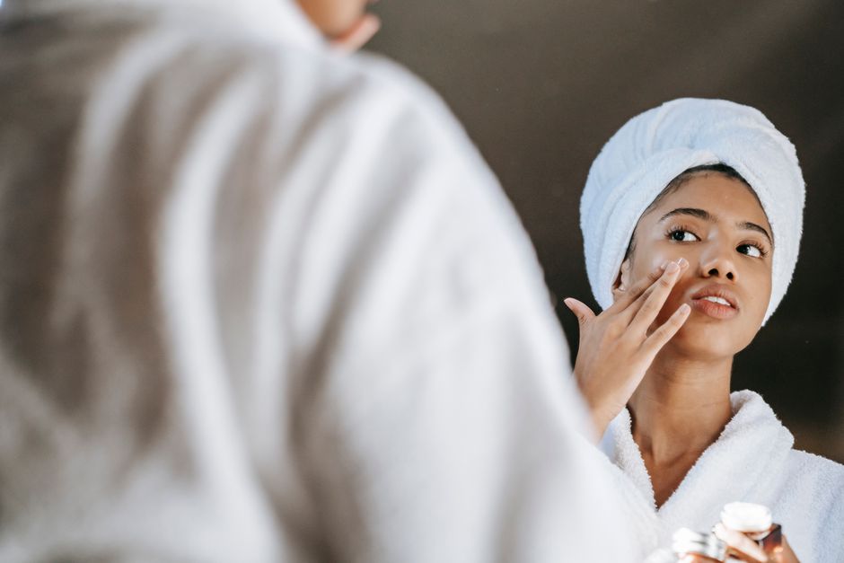 Back view of crop young ethnic lady in robe and terry towel applying cream on cheek while reflecting in mirror