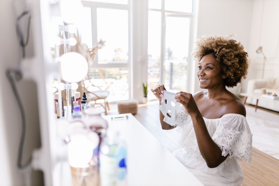Smiling woman holding face mask in bright, airy room near a vanity mirror.