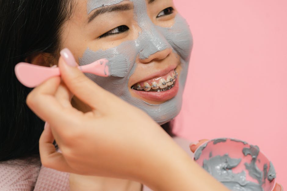 Close-up of a woman with braces applying a clay face mask, smiling cheerfully against a pink background.