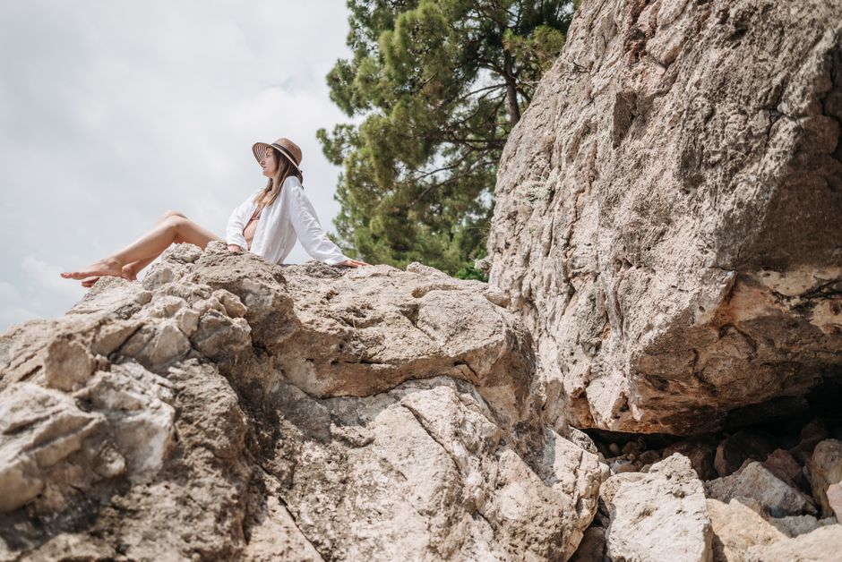 A woman in white long sleeves and hat relaxes on a rock with a scenic view.