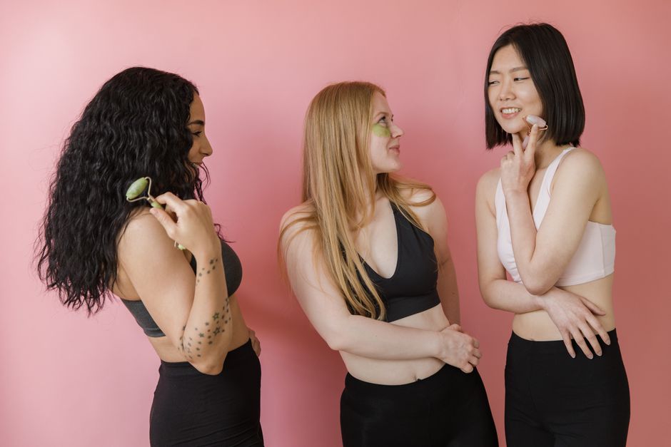 Three diverse young women enjoying a skincare routine with jade rollers against a pink backdrop.