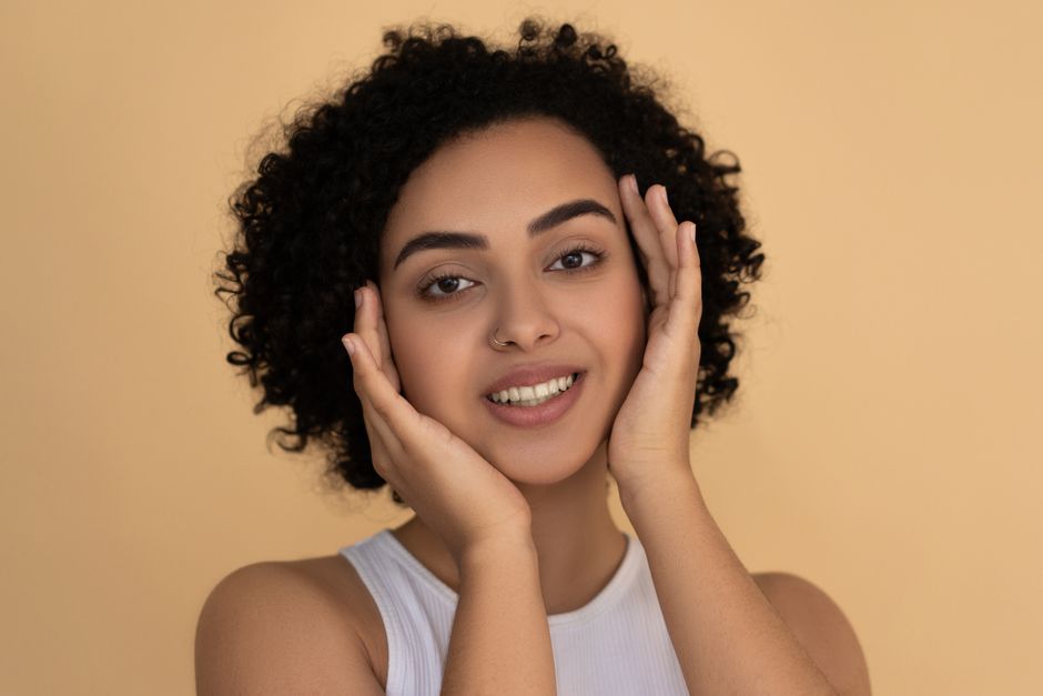 Close-up portrait of a woman with curly hair smiling and touching her face, exuding natural beauty.