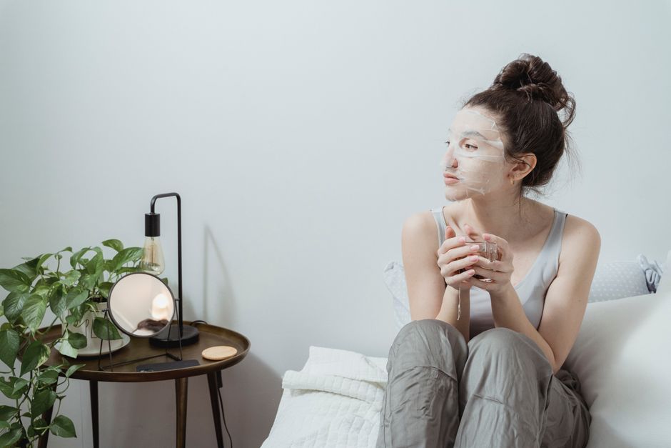 Woman sitting on bed with facial mask, enjoying skincare in a cozy indoor setup with plants.