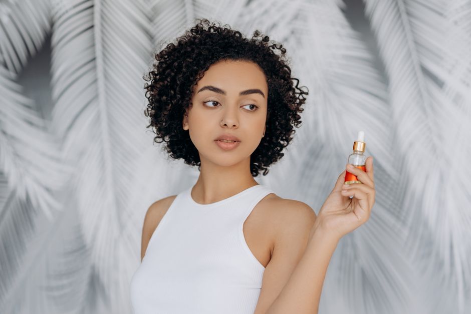 Young woman holding essential oil bottle with a serene expression against a white leafy background.