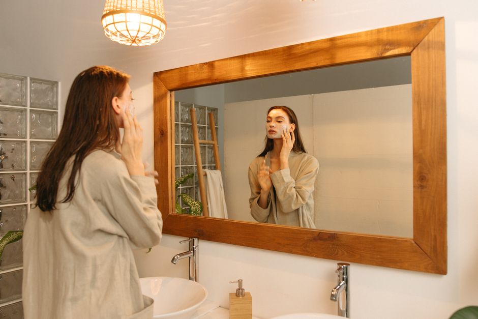 Woman applying skincare cream in front of a mirror in a cozy bathroom setting.