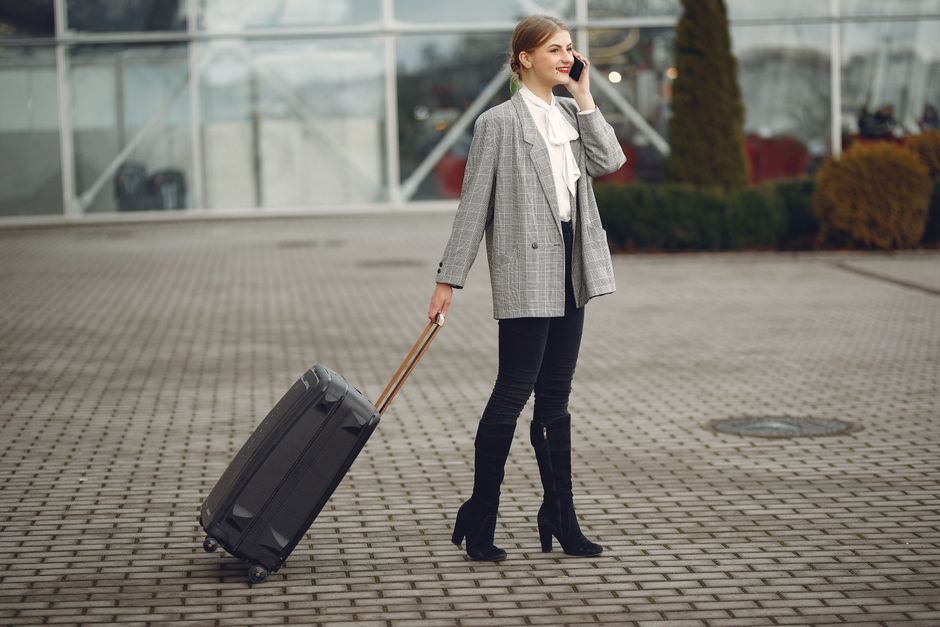 Full body of elegant female tourist in stylish clothes standing with suitcase and calling taxi by phone after arrival in modern city